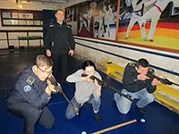 Трое людей целятся из винтовок, позади стоит мужчина в темной форме, на стене плакаты с борцами