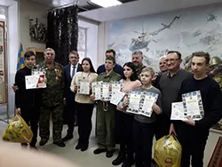 Взрослые и подростки стоят в музейной комнате и держат в руках дипломы, на стене рисунок с вертолетом