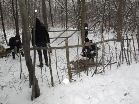 Люди в зимней одежде возле каркаса из жердей и веток в заснеженном лесу