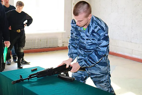Юноша в камуфляже склонился над автоматом на столе с зеленой скатертью, рядом стоят люди