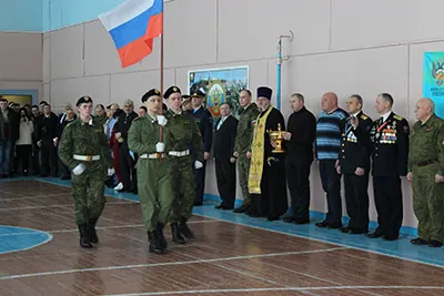 Люди в камуфляже и в форме стоят в спортзале, слева флаг России, рядом священник с кубком