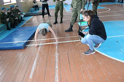 Девушка отжимается на полу спортзала, рядом стоят женщина с планшетом и человек в камуфляже