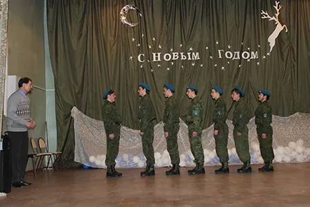Семеро ребят в камуфляже и голубых беретах стоят в шеренге на сцене, слева стоит мужчина