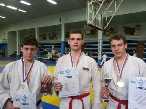 Три спортсмена в кимоно с медалями на лентах и грамотами в руках стоят в спортивном зале, у одного кубок