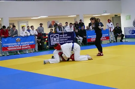 Двое бойцов в белых кимоно и красных шлемах борются на ковре, рядом судья, на заднем плане зрители
