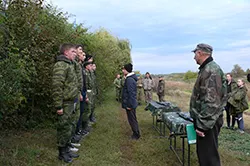 Люди в камуфляже стоят на поляне у кустов, рядом стоят столы