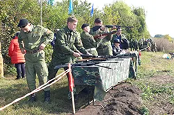 Люди в камуфляже у стола с оружием на открытой площадке, на заднем плане флаги
