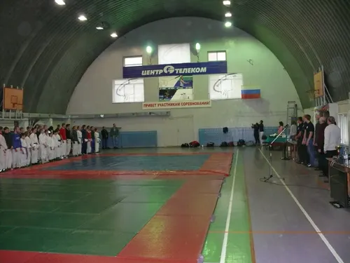 Спортсмены в белой форме стоят в шеренге в большом спортивном зале с арочным потолком